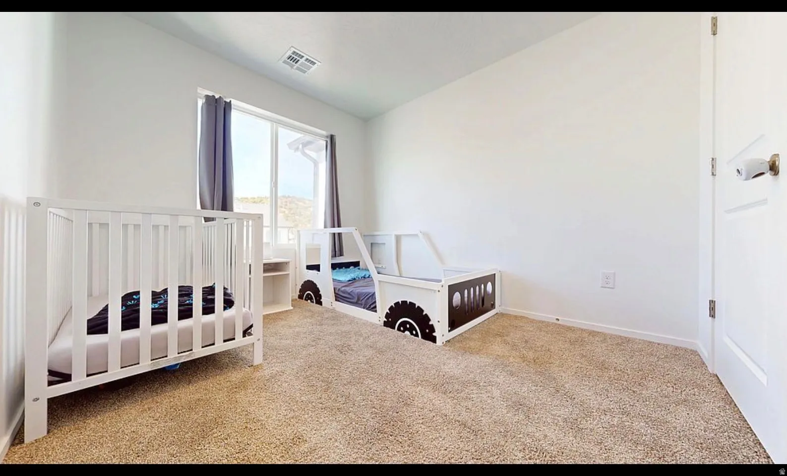 Carpeted bedroom with a large window, white walls, and a white door with bronze hardware