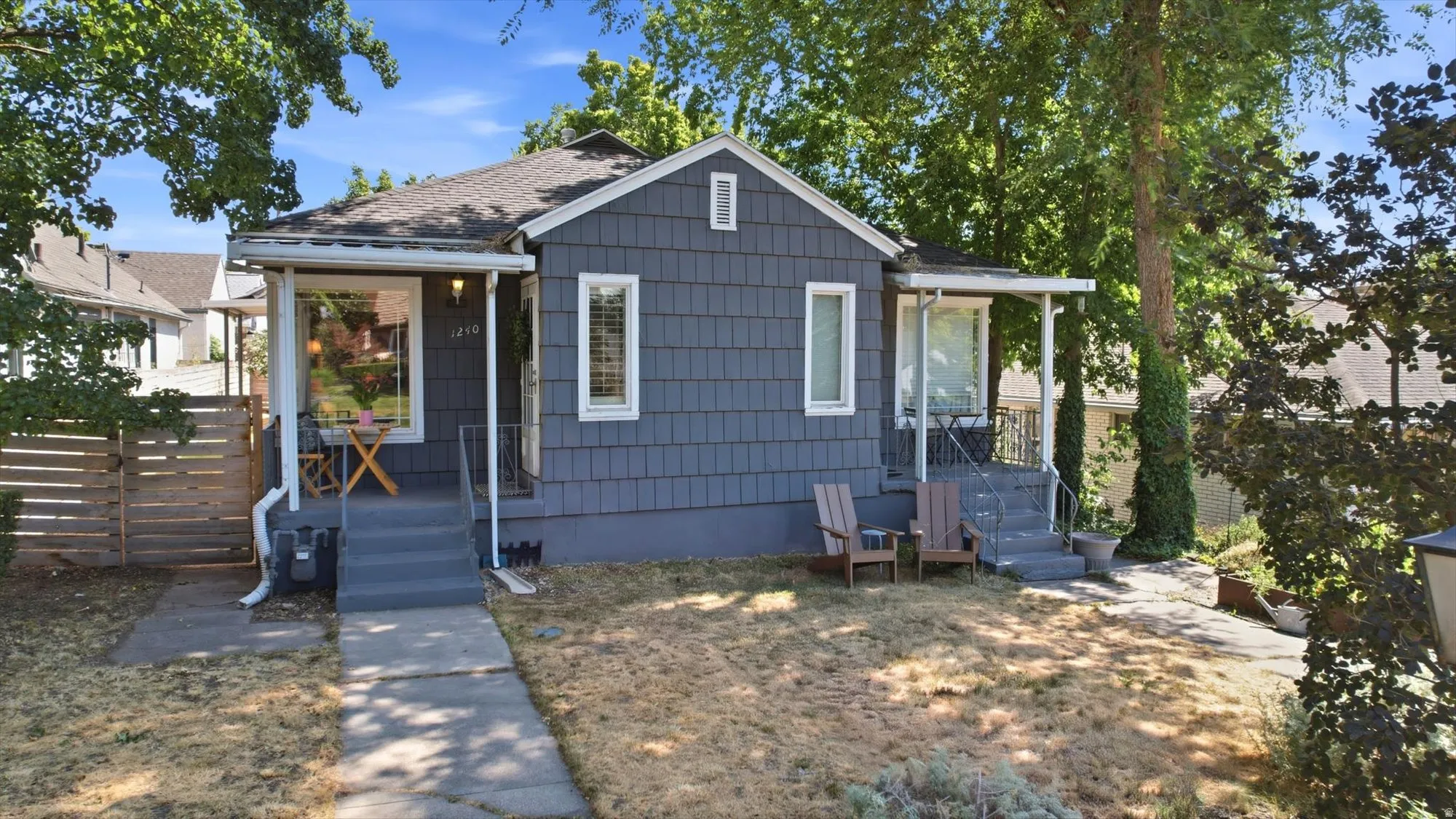 Shingle siding exterior with a covered front porch, concrete pathway, and mature trees