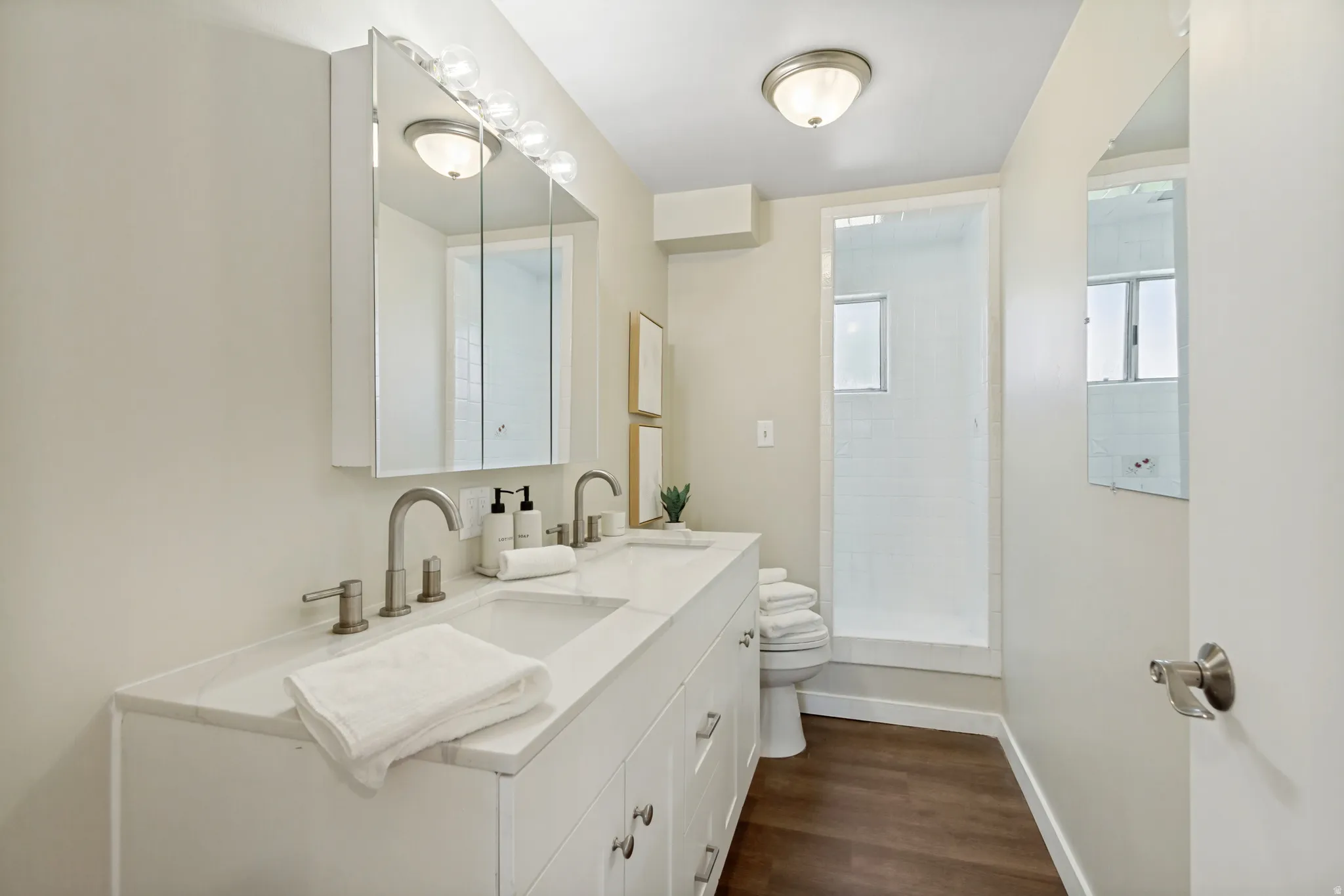 Bathroom featuring a dual vanity with undermount sinks, a shower enclosure with white subway tile, wood-finish flooring, and a recessed medicine cabinet with integrated lighting