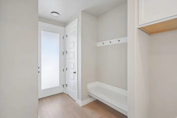 Entryway featuring a white frosted glass door, light wood-finish flooring, built-in bench seating, storage hooks, and overhead lighting