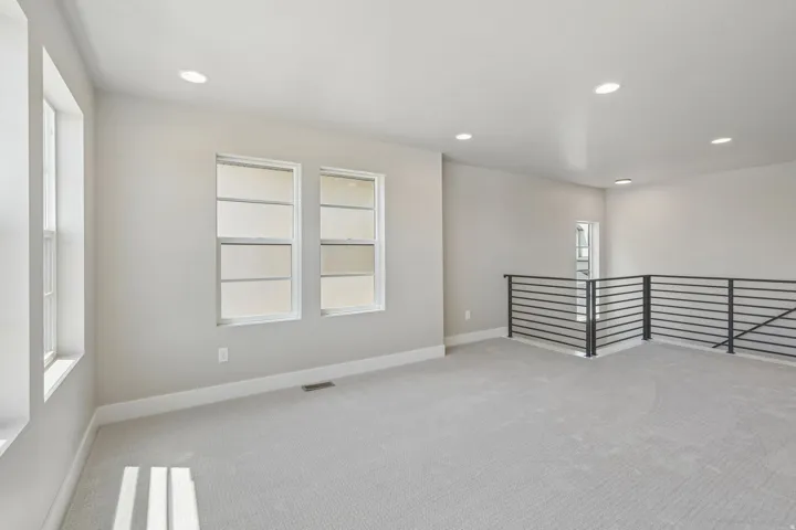 Spacious loft area featuring light gray carpeting, multiple windows, recessed lighting, and a modern horizontal bar railing system