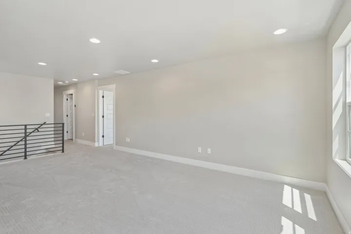 Spacious loft area featuring light gray carpeting, recessed ceiling lighting, a large window, white trim, and a black metal handrail