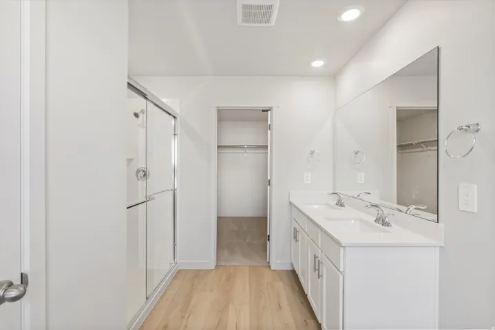 Bathroom featuring a dual vanity with white cabinetry and chrome fixtures, a frameless glass shower enclosure, wood-finish flooring, and recessed lighting