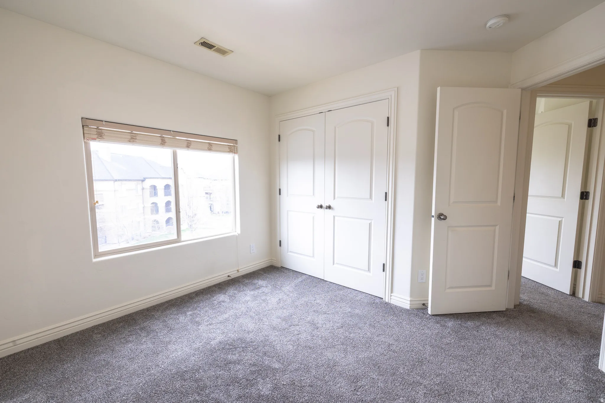 Bright room featuring neutral-toned walls, gray carpeting, a window with horizontal blinds, and a double-door closet with arc-top panels