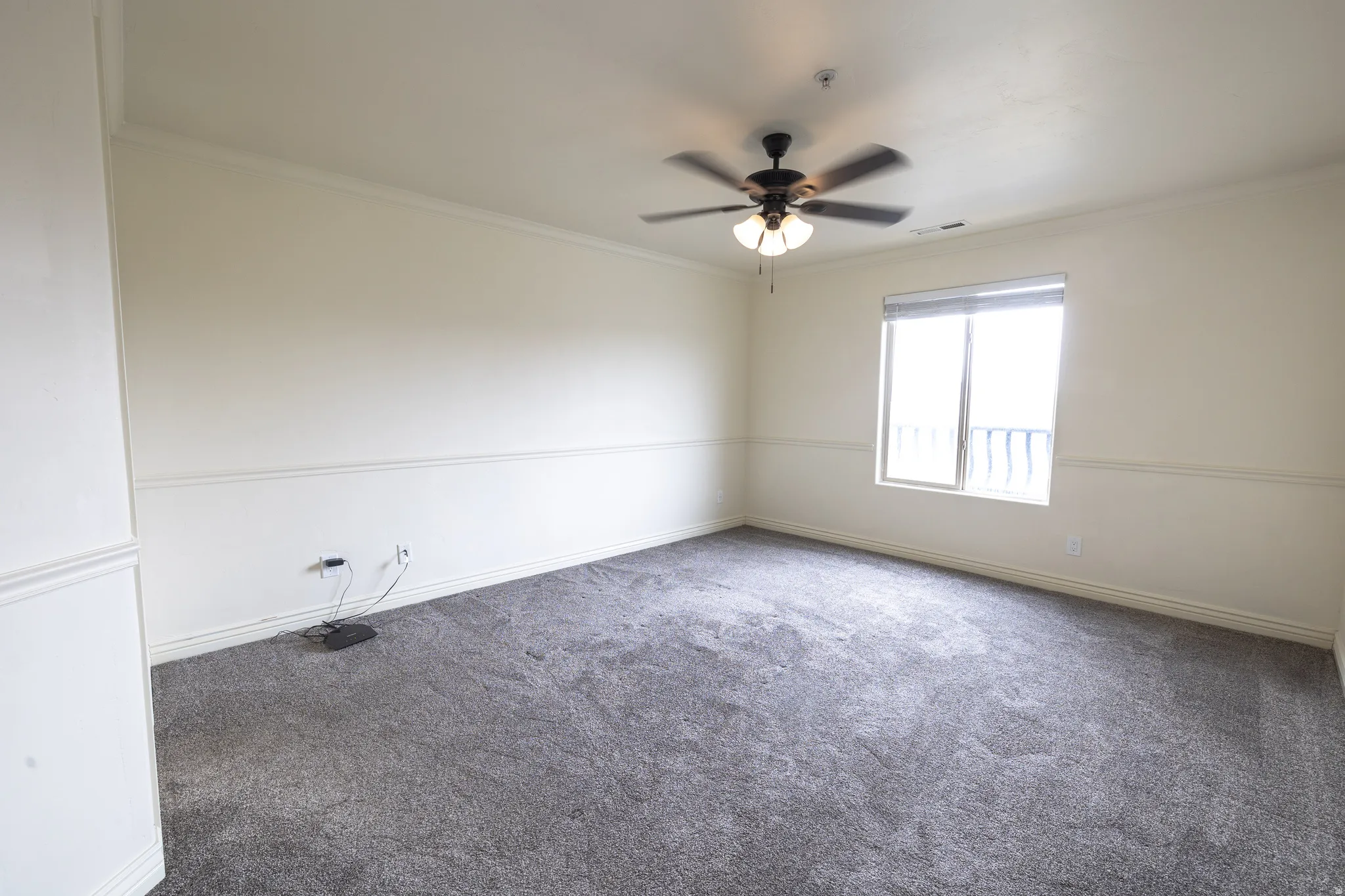 Spacious room featuring gray carpeting, a window with natural light, crown molding, a ceiling fan with light fixture, and wainscoting details