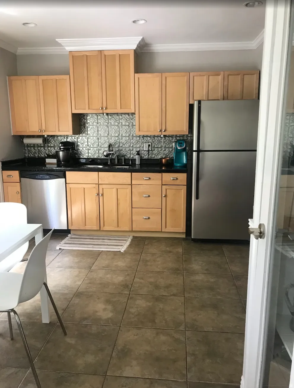 Kitchen featuring light wood cabinetry, black countertops, a metallic tile backsplash, stainless steel appliances, and large format floor tiles
