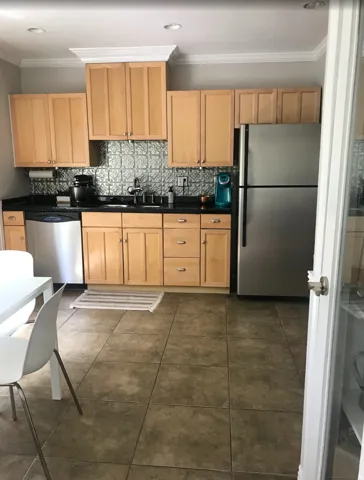 Kitchen featuring light wood cabinetry, black countertops, a metallic tile backsplash, stainless steel appliances, and large format floor tiles