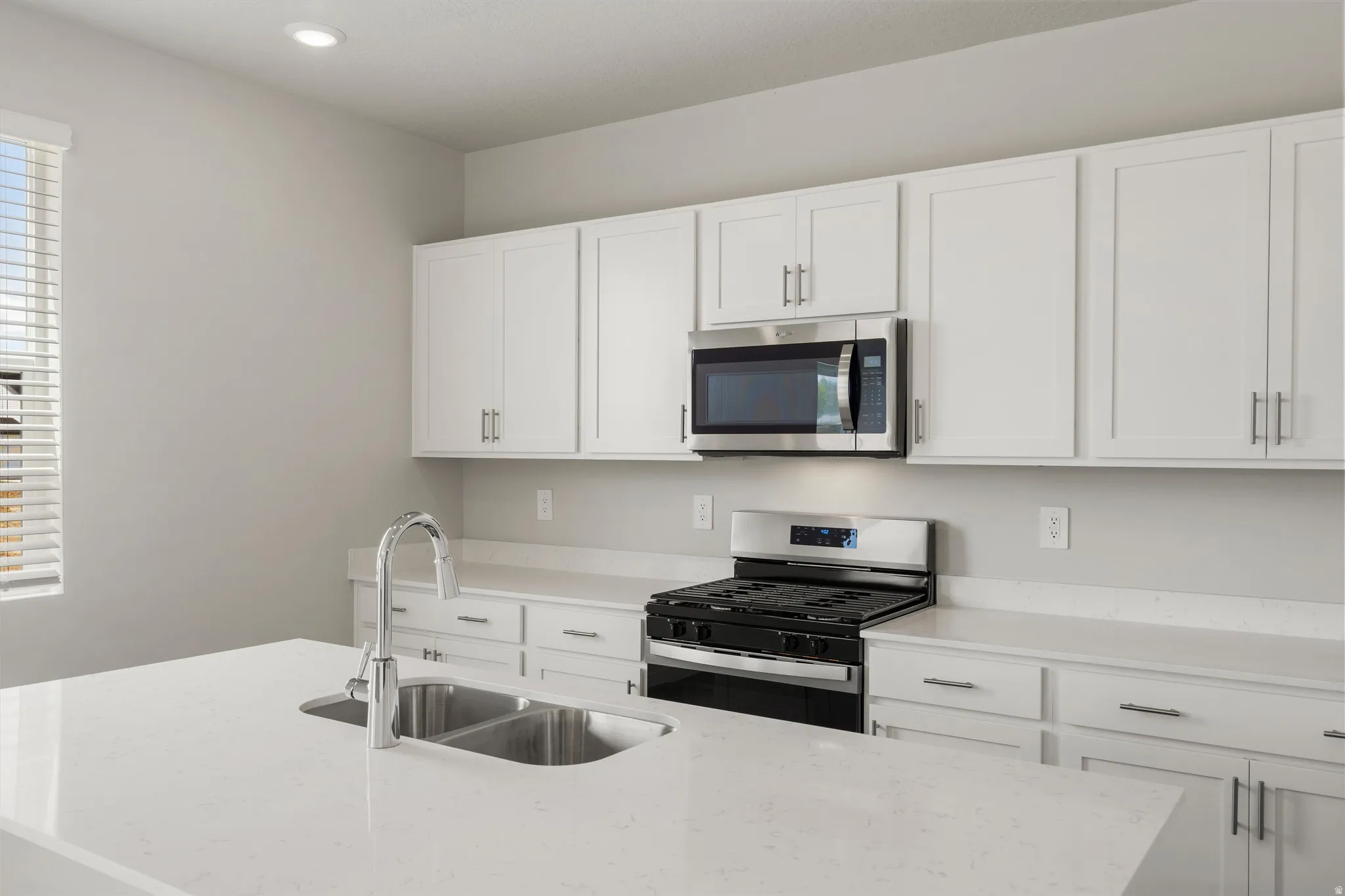 Kitchen featuring a large central island with a double basin sink and a high-arc faucet, white shaker-style cabinetry with brushed nickel hardware, light-toned countertops, and stainless steel appliances including a range and over-the-range microwave
