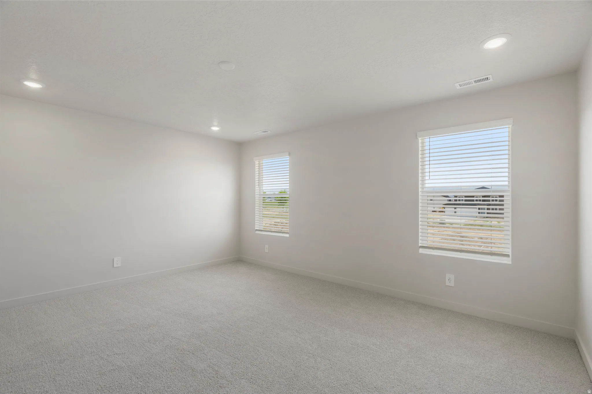 Spacious room featuring neutral carpeting, light gray walls, two windows with blinds, and recessed lighting