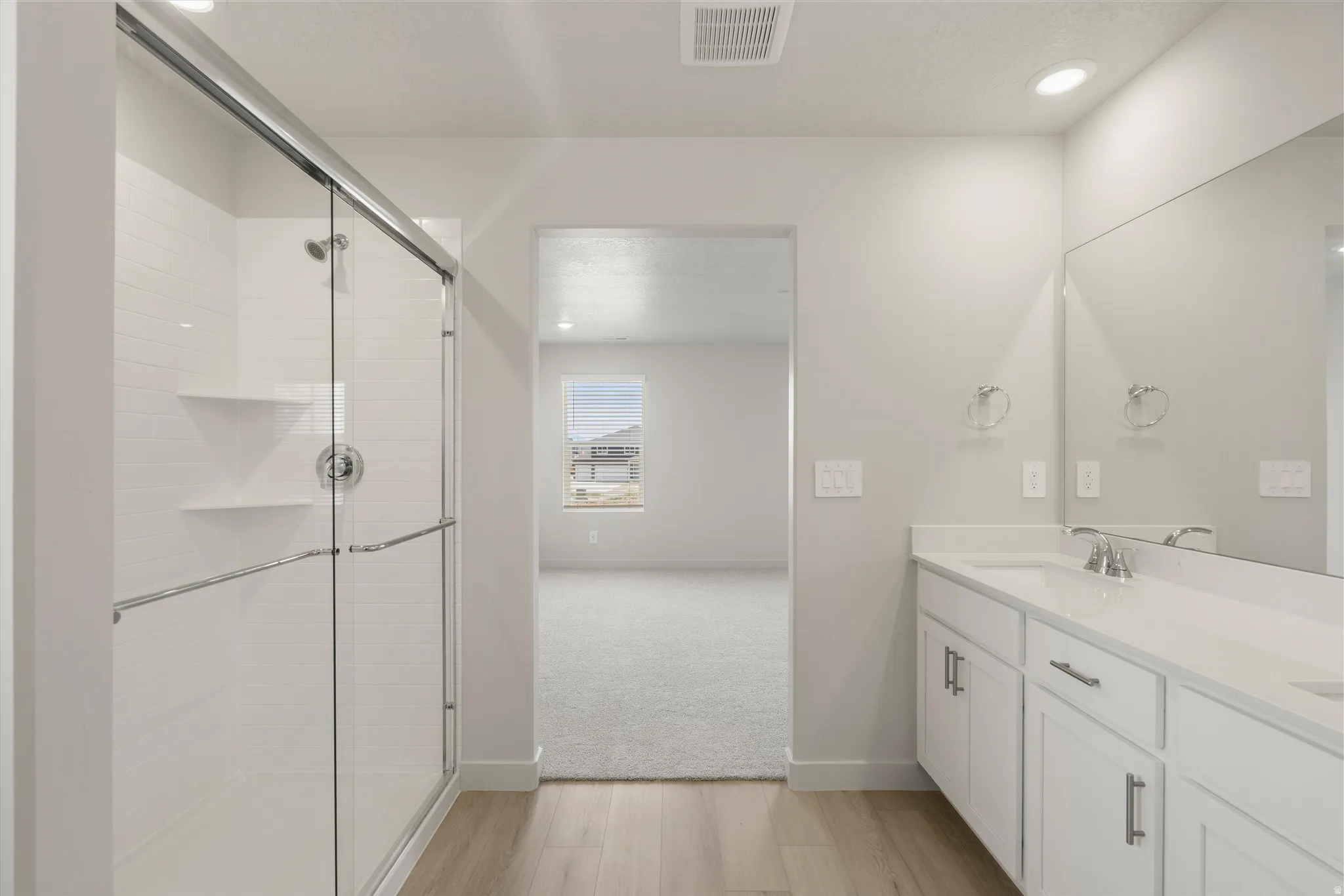Bathroom featuring wood-finish flooring, a double vanity with white cabinetry and integrated sinks, a large wall-mounted mirror, and a glass-enclosed shower with white tiled walls