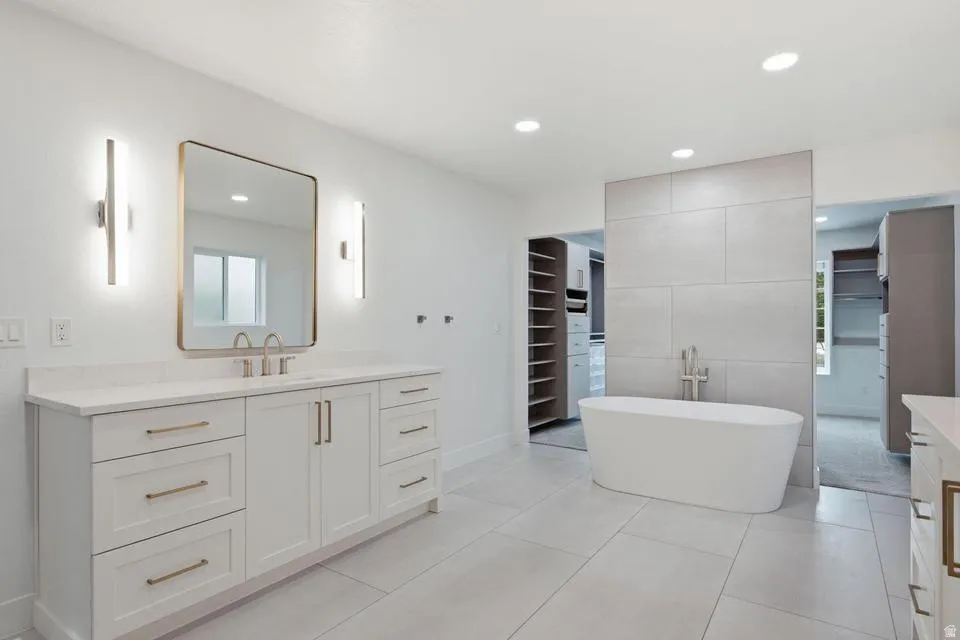 Spacious bathroom featuring a freestanding tub, large format floor tiles, dual vanity with shaker cabinetry, gold-tone hardware, and integrated lighting