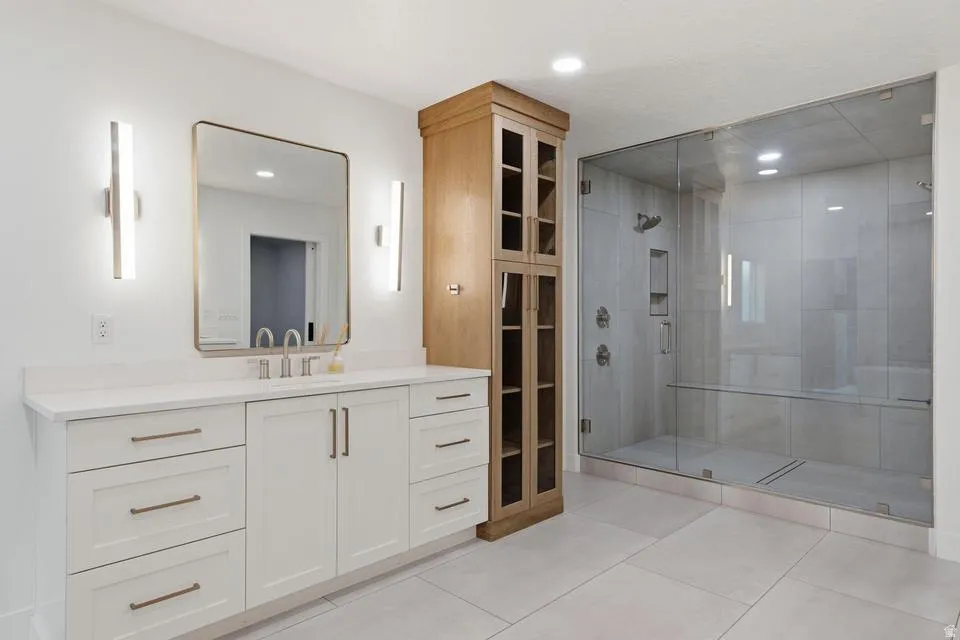 Spacious bathroom featuring a frameless glass shower enclosure with built-in bench, a double vanity with white cabinetry and quartz countertop, a large framed mirror, and a tall built-in wood-finish linen cabinet
