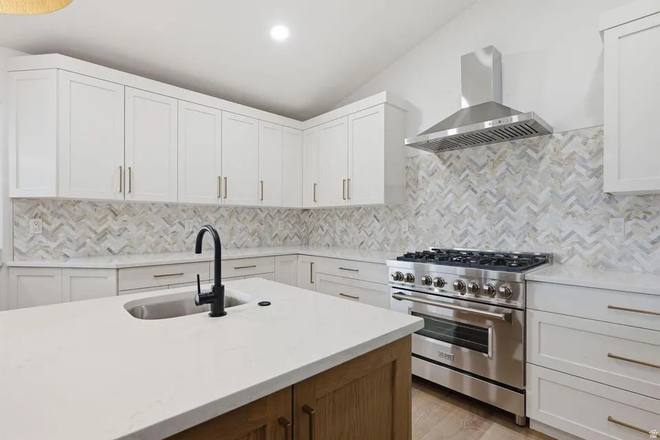 Gourmet kitchen featuring a sizable central island with an undermount sink and matte black faucet, white shaker cabinetry with gold-tone hardware, a stainless steel range with a coordinating hood, light-toned wood-finish flooring, and a unique herringbone