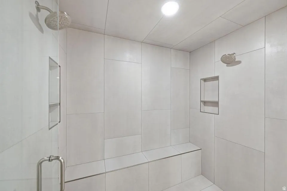 Spacious walk-in shower featuring large format light-toned tile, dual showerheads, built-in bench seating, and two recessed shower niches