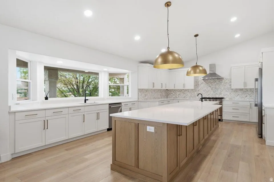 Open-concept kitchen featuring an expansive central island with a light wood-finish base, quartz countertops, white cabinetry with contemporary hardware, a herringbone tile backsplash, and wood-finish flooring