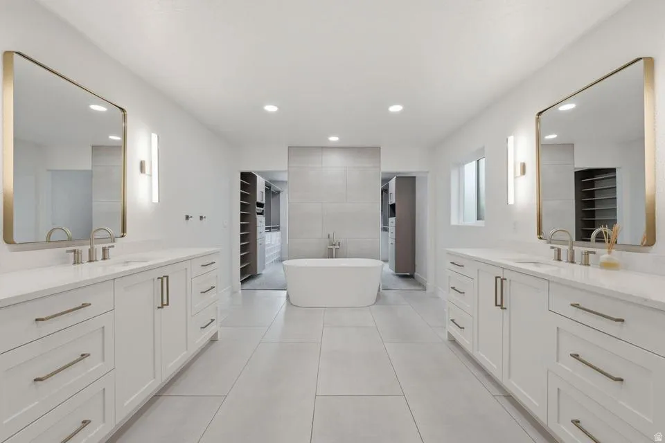 Spacious bathroom featuring a freestanding tub, dual vanities with white cabinetry, gold-framed mirrors, and large format tile flooring