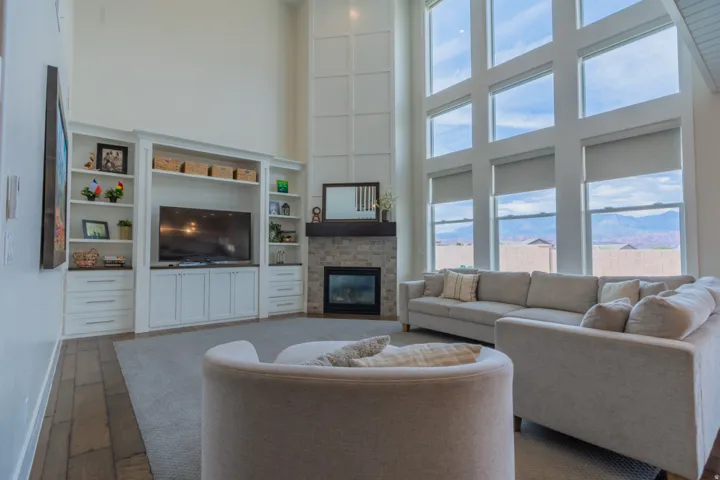 Living area featuring a two-story wall of windows, built-in entertainment center with shelving and storage, a stacked stone fireplace with a dark wood mantel, and tall ceilings with detailed wall paneling