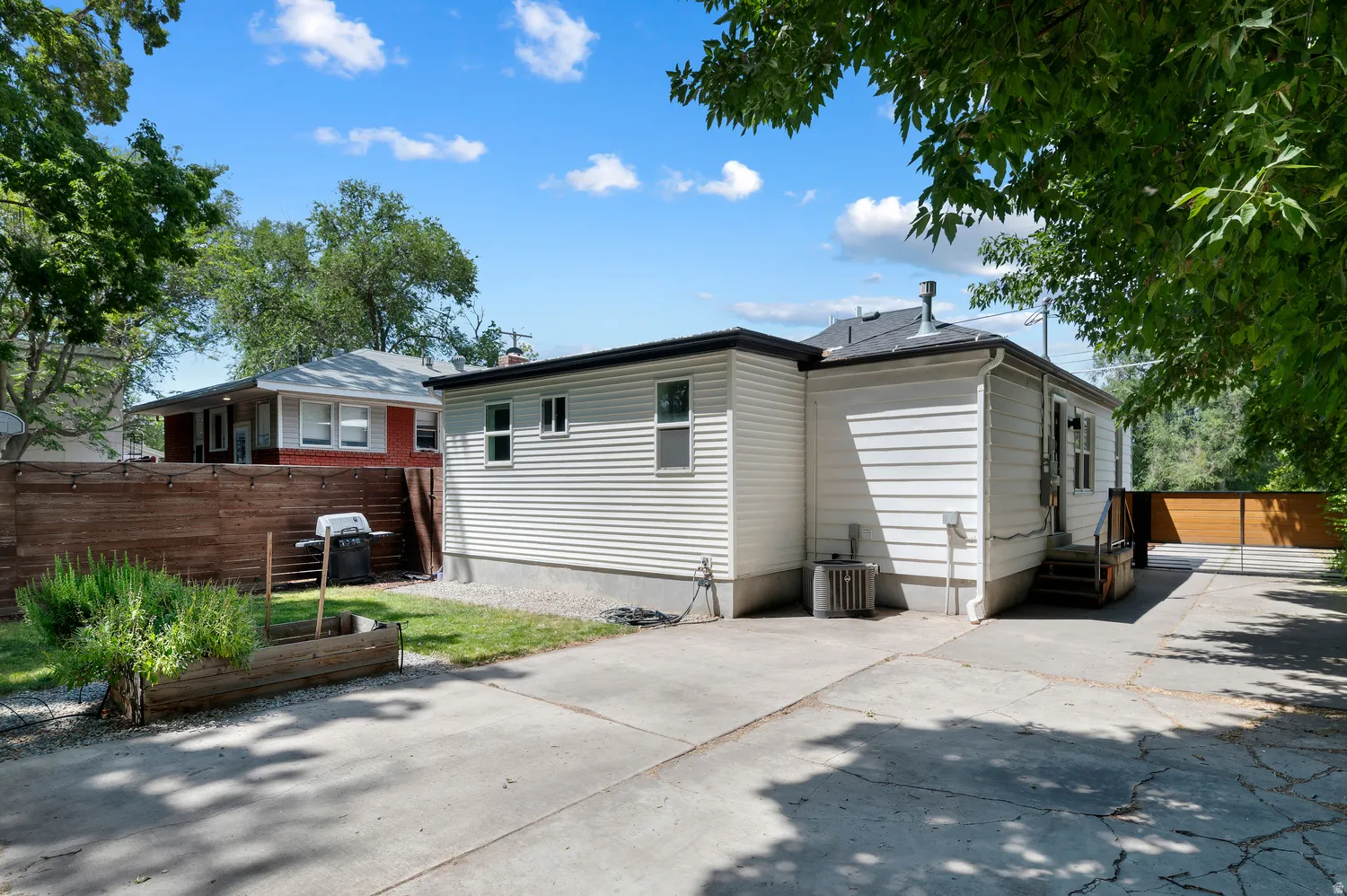 Rear exterior featuring white horizontal siding, a concrete driveway, a wooden privacy fence, a small grassy area, and mature perimeter trees