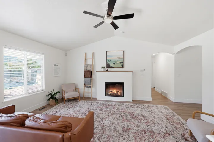 Vaulted ceiling living space featuring a built-in fireplace with wood-finish mantel, wide window with blinds, ceiling fan, and wood-finish flooring