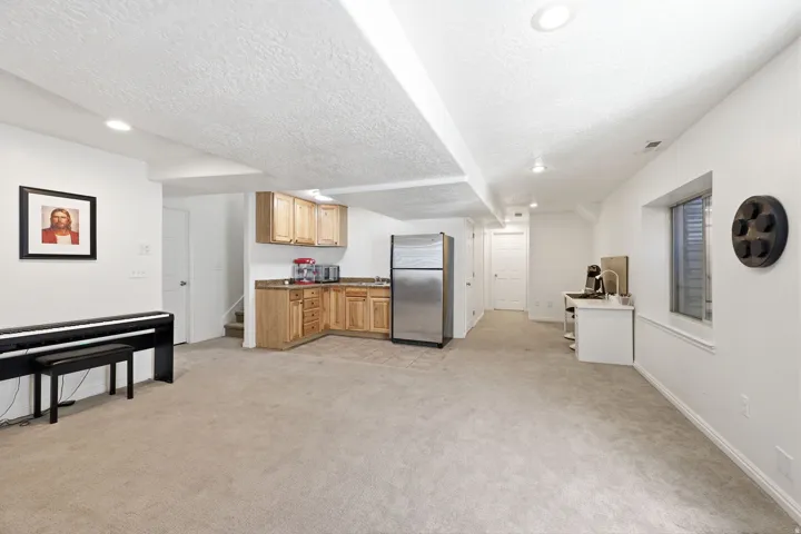 Spacious finished area featuring recessed lighting, light-toned carpet, a kitchenette with wood cabinetry and a stainless steel refrigerator, and a dedicated workspace
