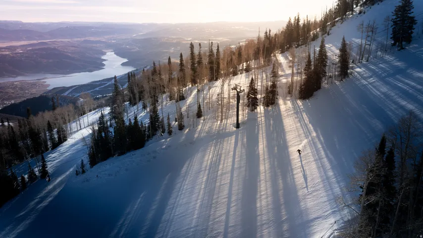 Snow-covered mountain slope featuring evergreen trees and a ski lift structure, with a distant body of water and valley visible under a bright sky