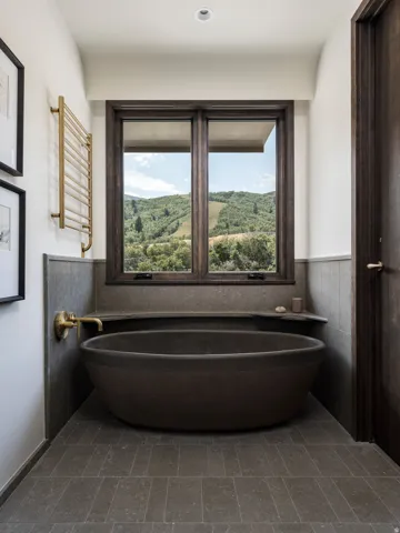 Freestanding soaking tub with dark stone finish, featuring brass wall-mounted faucet and brass towel warmer