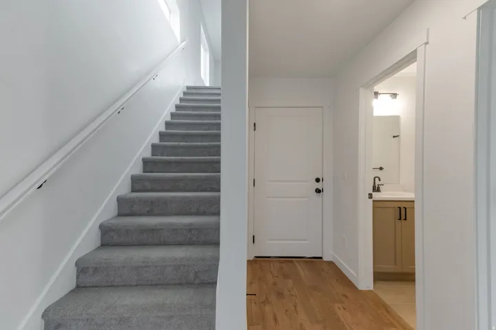 Carpeted staircase with white handrail and upper windows, complemented by wood-finish flooring, a white paneled door, and a bathroom entry featuring a vanity with a white countertop and dark hardware