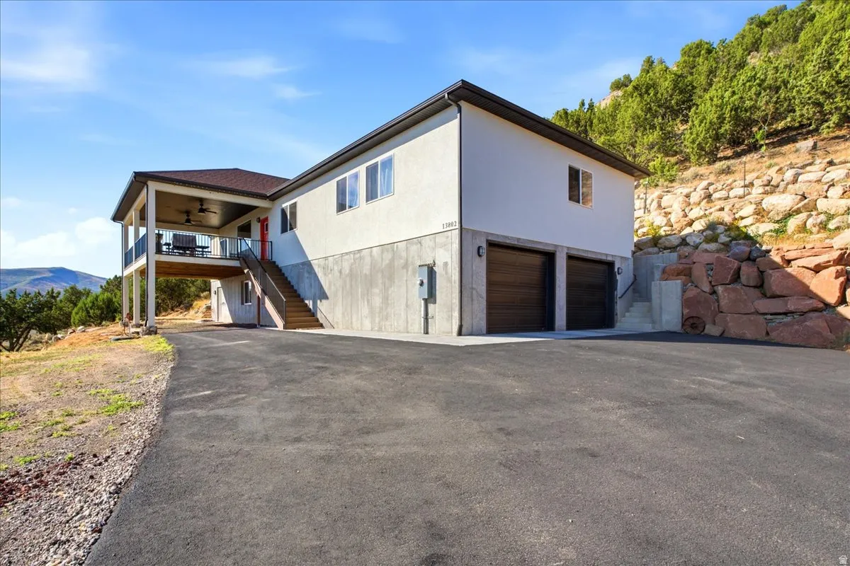 Contemporary two-story exterior with a covered balcony, lower-level garage with two dark wood-finish doors, a paved driveway, and natural rock landscaping