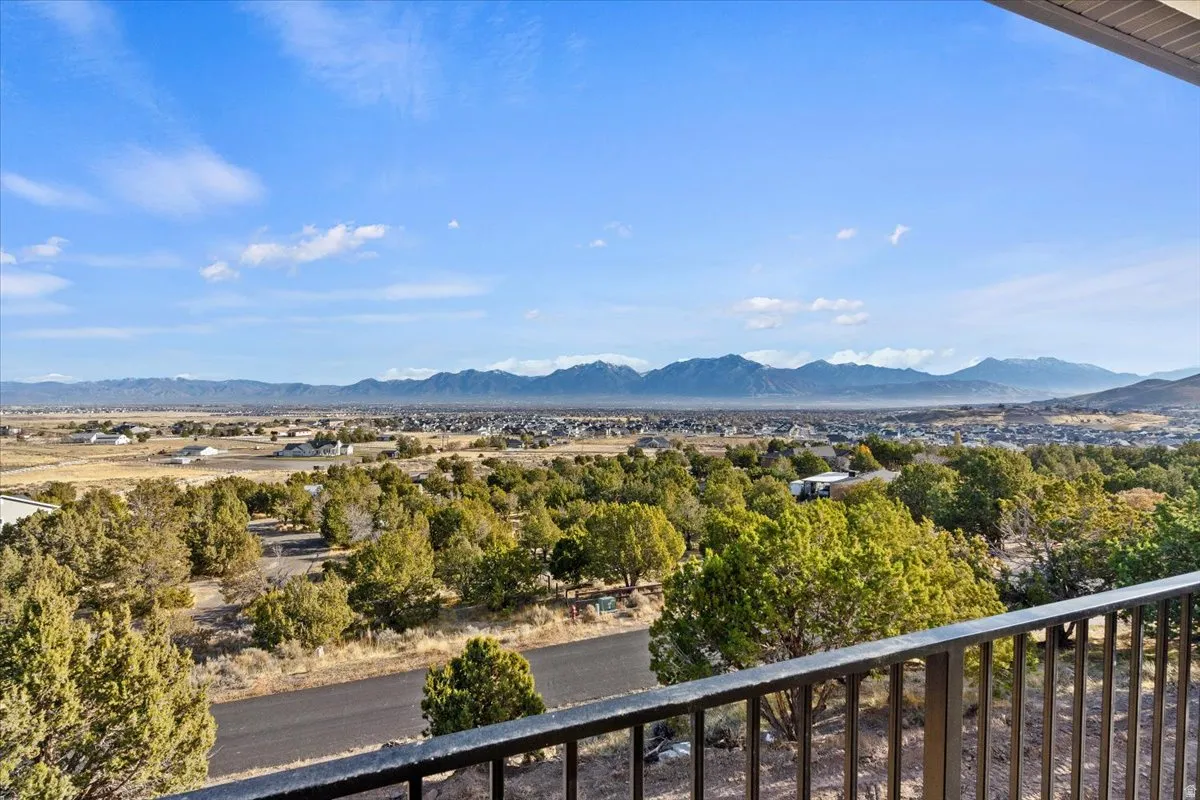Expansive mountain and valley scenery from a private balcony, featuring a black metal railing, mature evergreen landscaping, and a distant residential community