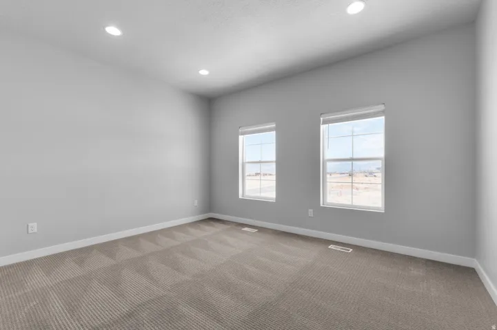Recessed lighting illuminates a spacious room featuring two large windows, neutral gray walls, and patterned carpet flooring