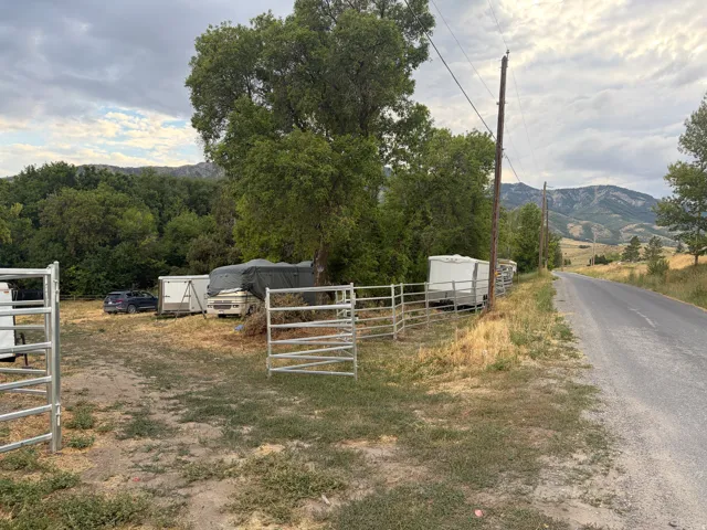 Gated property entrance opening to a gravel lot, featuring mature trees and a mountain backdrop