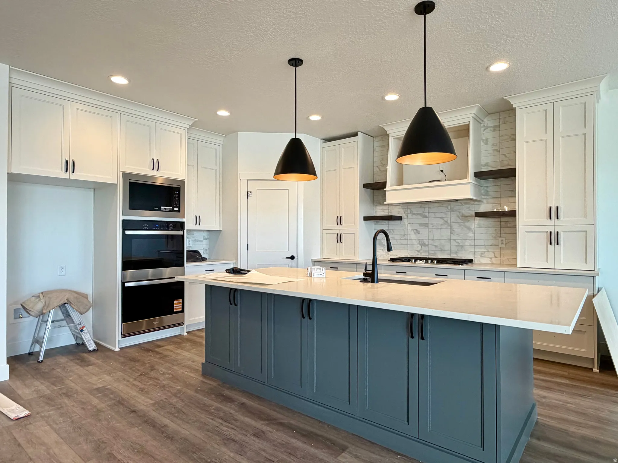 Kitchen featuring a substantial island with a light-toned countertop, white shaker-style cabinetry, stainless steel built-in appliances, a subway tile backsplash, and wood-finish flooring