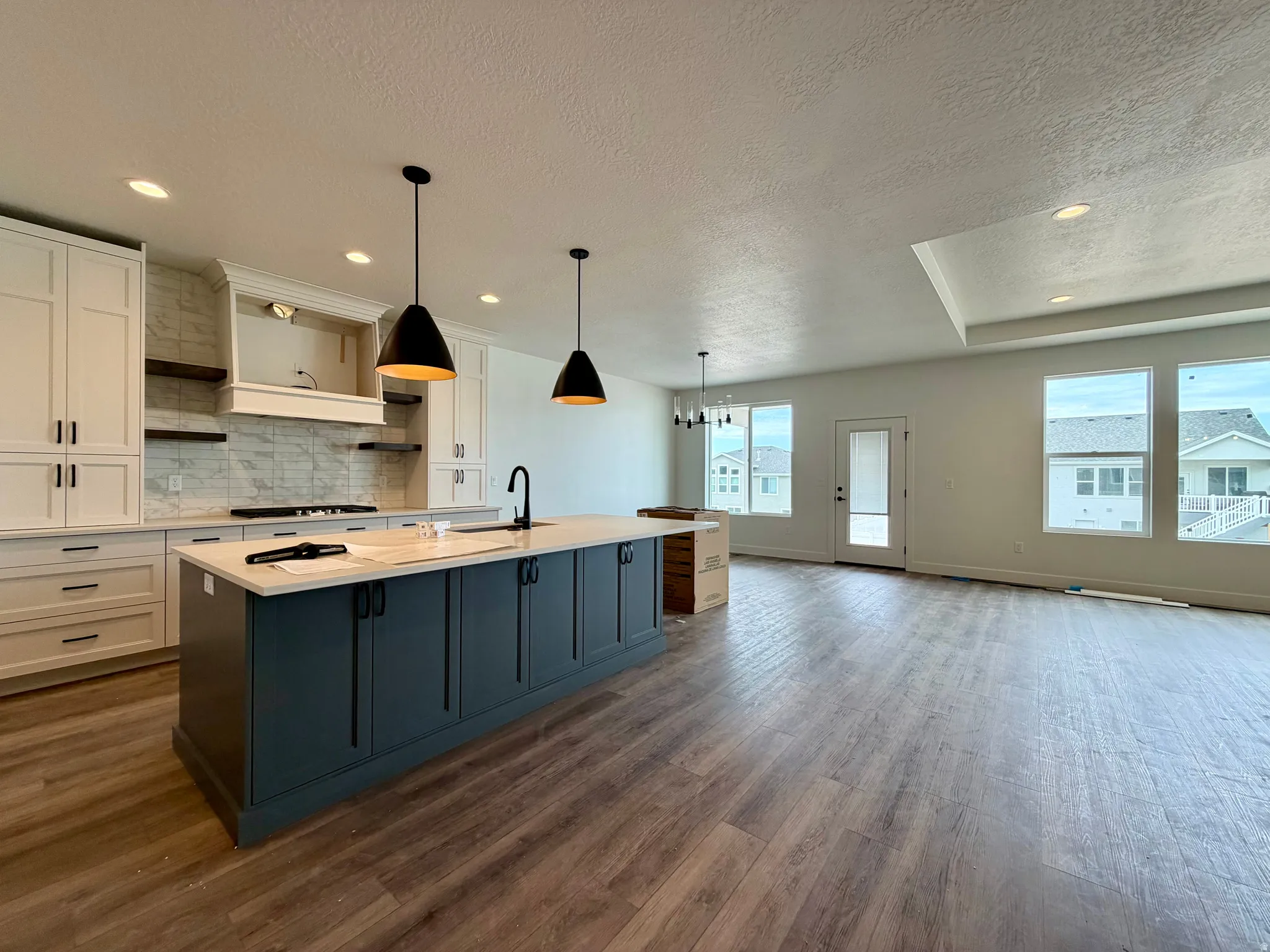 Spacious open-concept kitchen and living area featuring a large island with sink, white shaker cabinetry, wood-finish flooring, a tray ceiling, and multiple windows