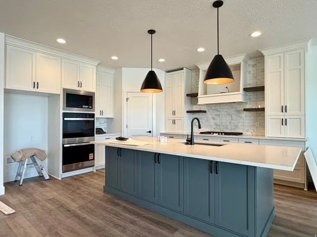 Kitchen featuring a substantial island with a light-toned countertop, white shaker-style cabinetry, stainless steel built-in appliances, a subway tile backsplash, and wood-finish flooring