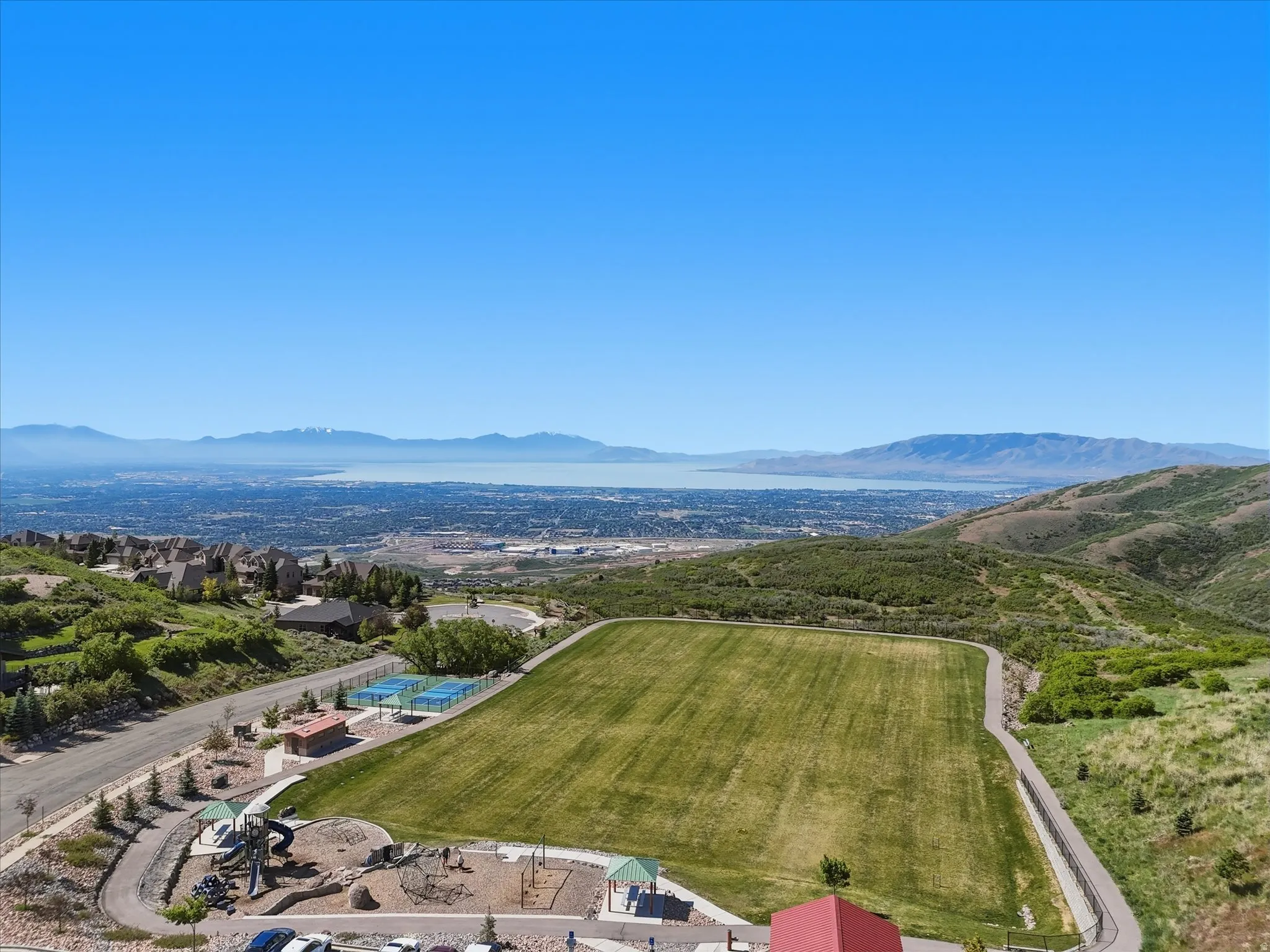 Expansive recreational field with surrounding walking path, two tennis courts, and a playground area, all set against a backdrop of distant mountains and a body of water
