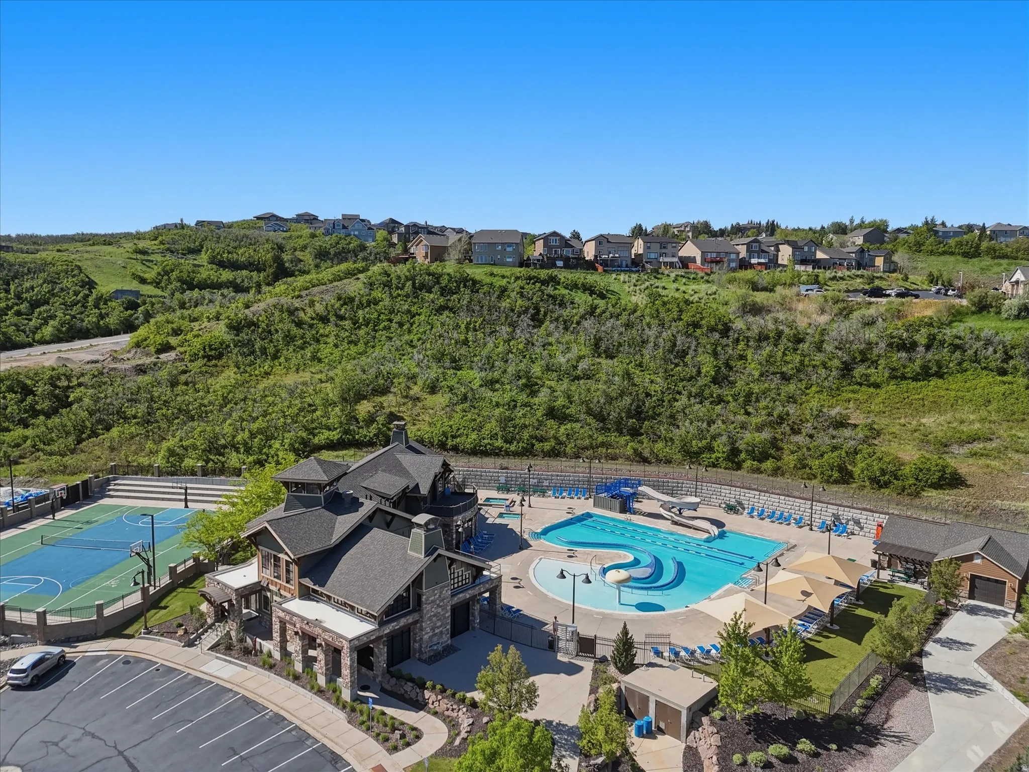 Community amenity center featuring a multi-sport court, a stone and siding clubhouse, multiple swimming pools, a waterslide, and shaded lounge areas