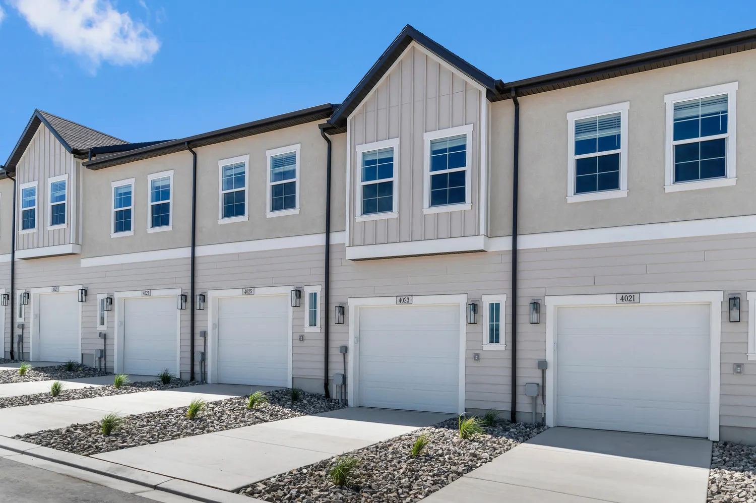Contemporary townhome exterior featuring light-colored siding, stucco accents, white-framed windows, individual garage doors, and exterior lighting fixtures