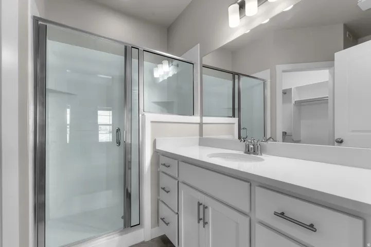 Walk-in shower with sliding glass door, vanity with dual sinks, expansive counter space, white cabinetry, and a large mirror