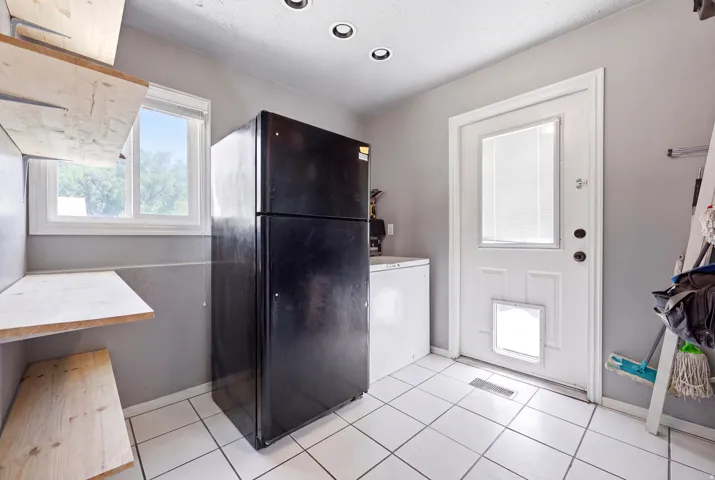 Utility room featuring white square tile flooring, a window with exterior views, built-in wooden shelving, a white paneled door with a glass insert, and recessed ceiling lighting