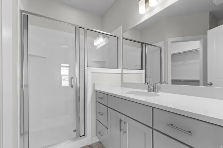 Glass-enclosed shower, single vanity with integrated sink, light gray cabinetry, wall-mounted light fixture, and wood-finish flooring