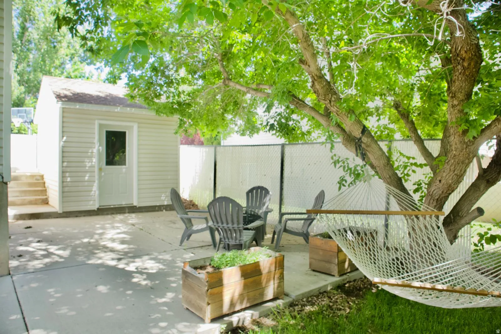 Concrete patio area featuring a detached storage shed with vinyl siding, mature shade tree, and wood planter boxes