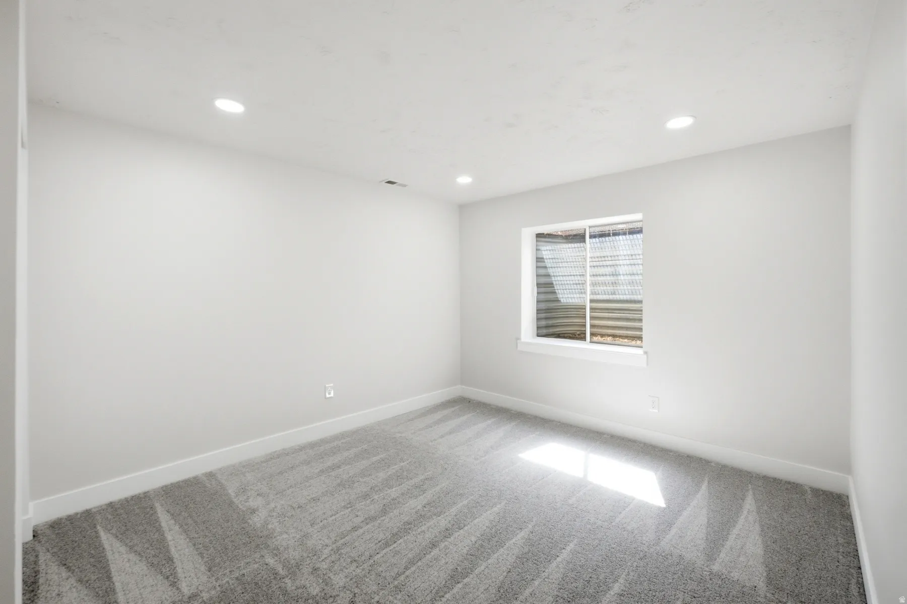 Spacious room featuring light gray walls, plush gray patterned carpeting, a white trim window, and recessed ceiling lighting