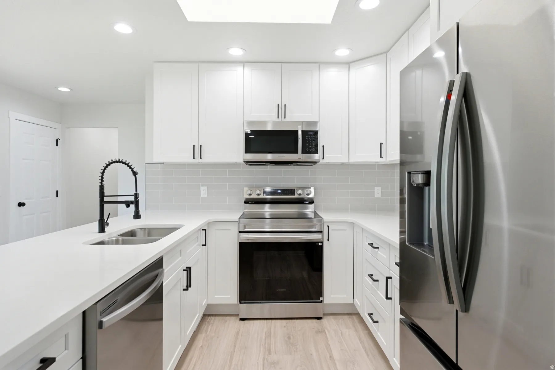 Modern kitchen featuring white cabinetry, light countertops, stainless steel appliances, a black gooseneck faucet, and light wood-finish flooring