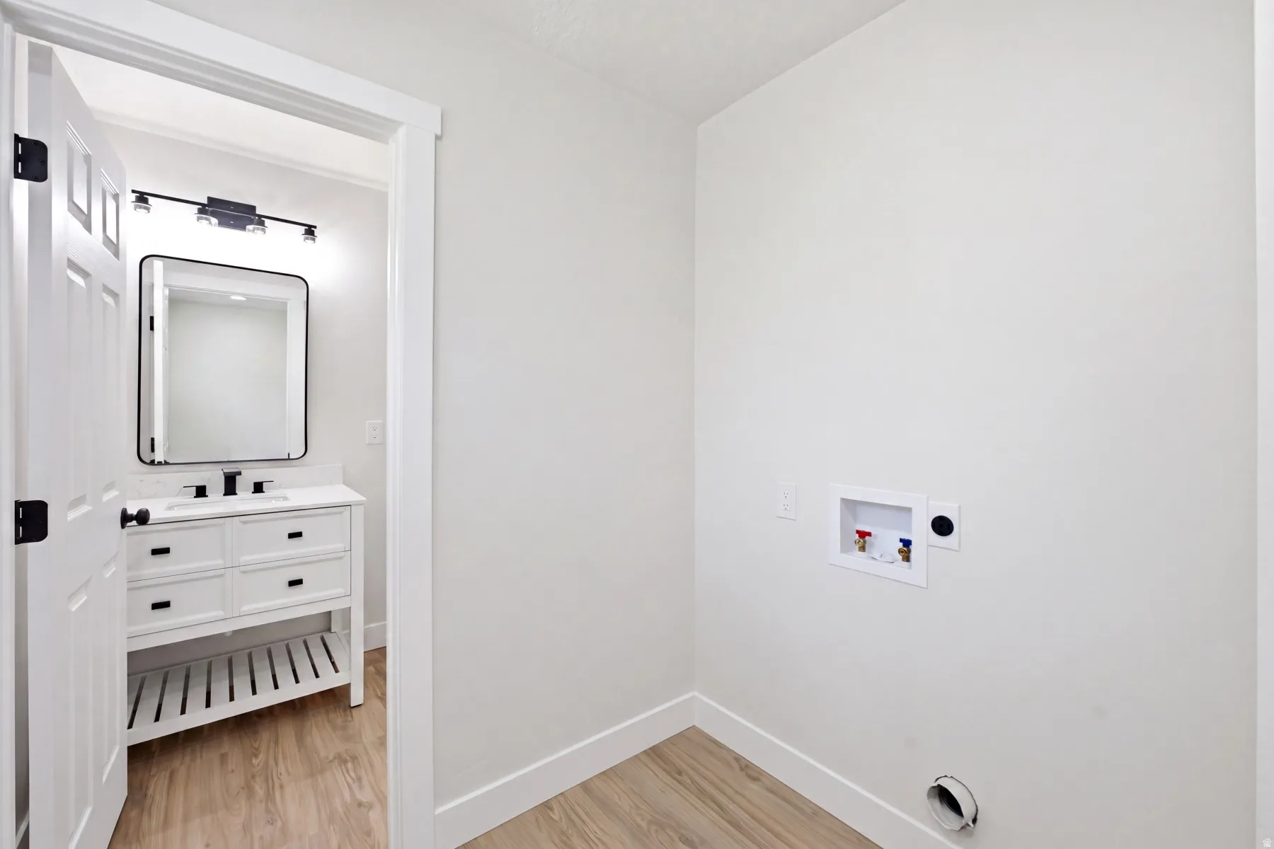 Laundry area with wood-finish flooring and a white vanity featuring a rectangular sink, black faucet, and modern black-framed mirror