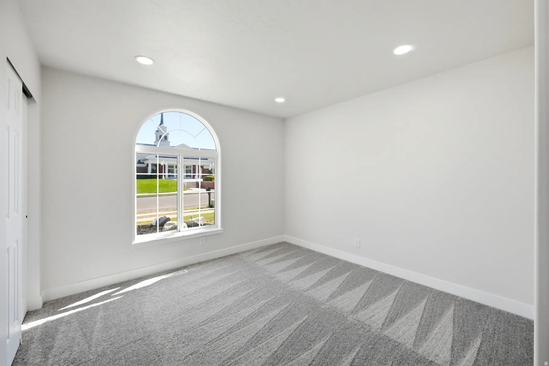 Spacious room featuring a large arched window with grid panes, recessed lighting, neutral wall tones, patterned carpet flooring, and a sliding two-panel closet door