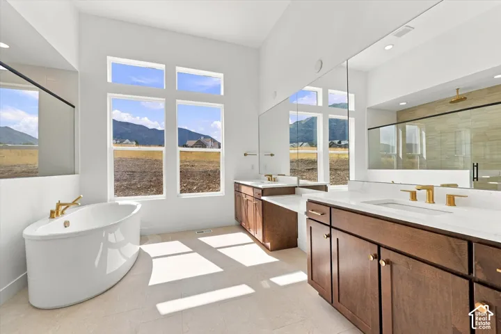 Freestanding soaking tub with brass fixtures, dual-sink vanity with light-colored stone countertops, large-format tile flooring, oversized mirrors, and walk-in glass-enclosed shower