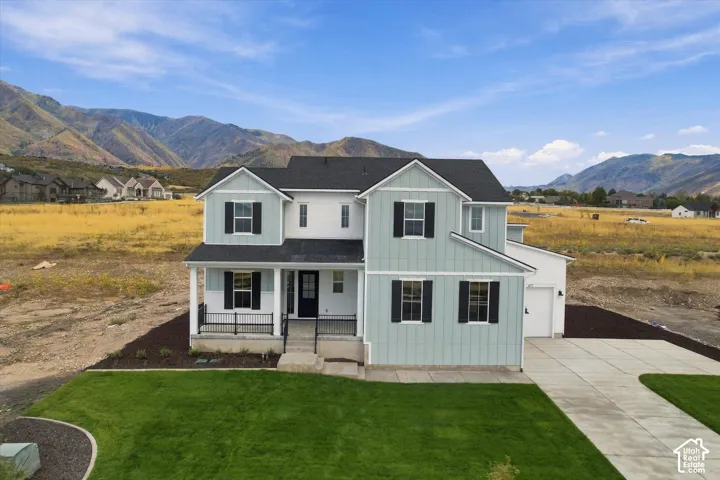 Two-story residence featuring board-and-batten siding, a covered front porch with metal railing, black shutter accents, and a concrete driveway, set against a mountain backdrop