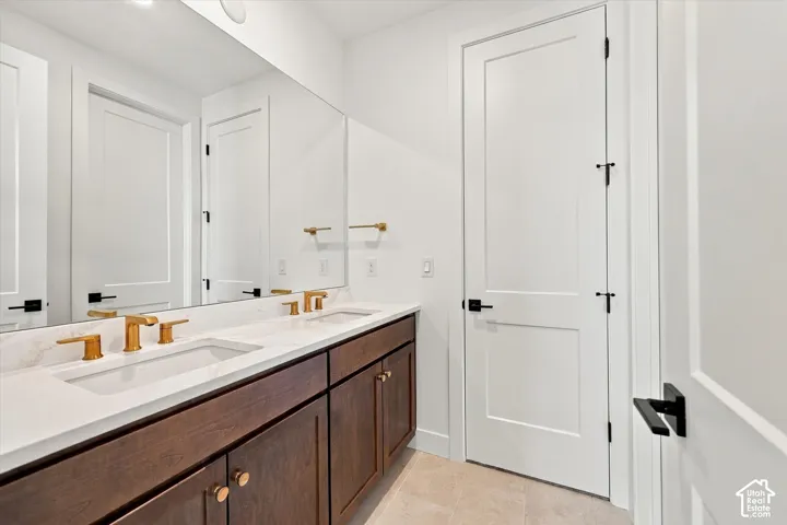 Double vanity featuring wood-finish cabinetry, white solid-surface countertop with dual under-mount sinks, gold-tone fixtures, and expansive frameless wall mirror
