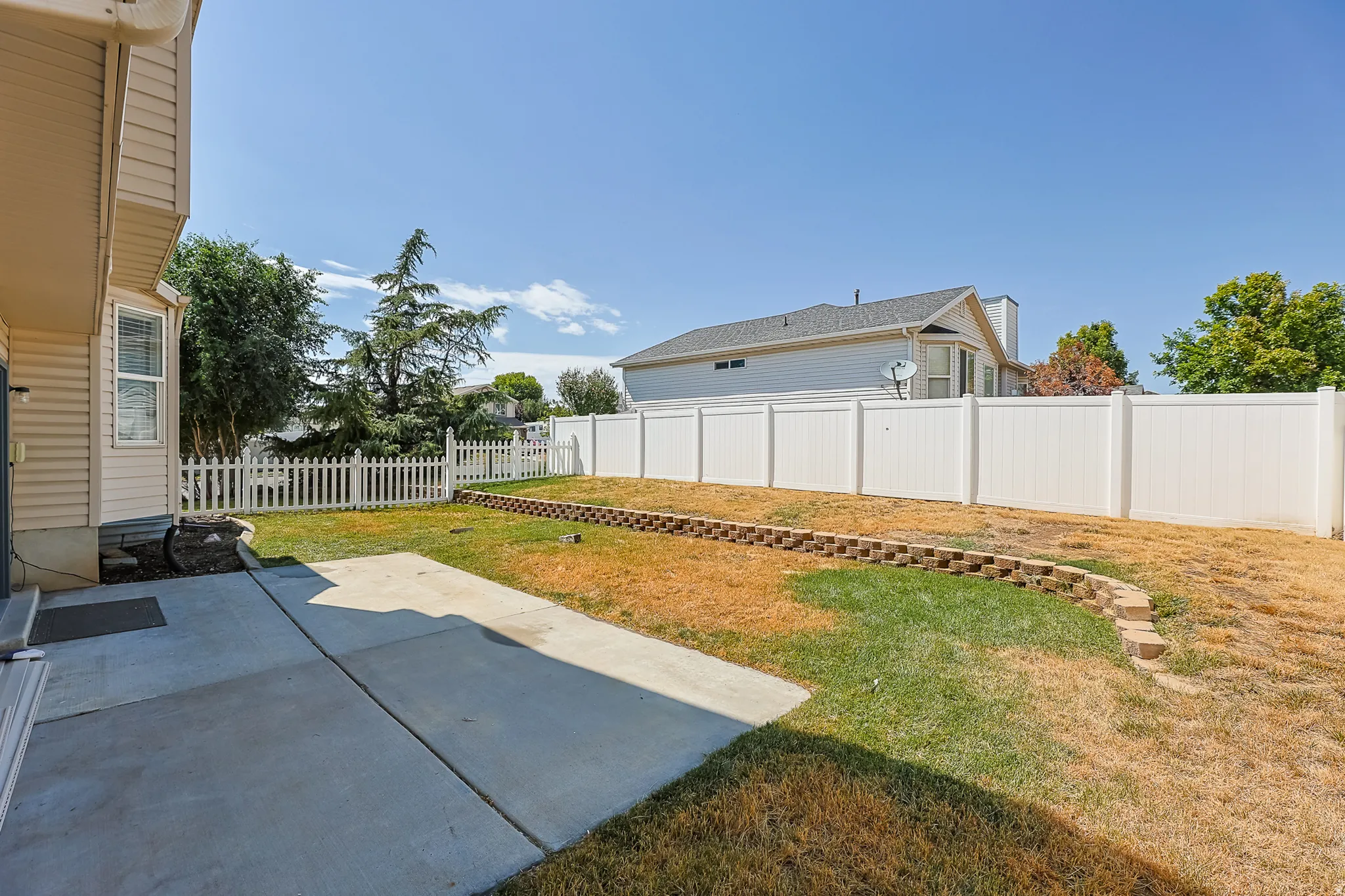 Spacious backyard featuring a concrete patio, white picket fence, privacy fencing, and a tiered retaining wall