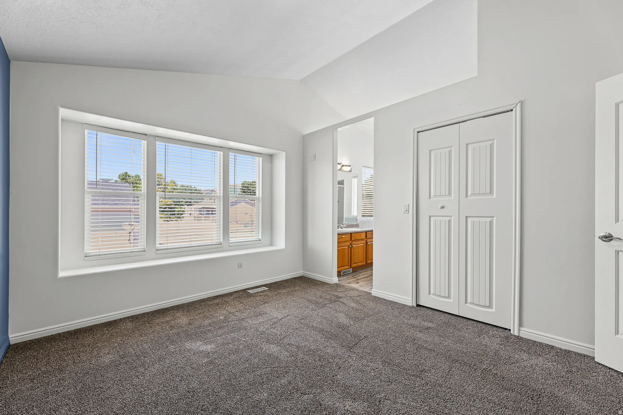 Spacious room featuring vaulted ceilings, a large three-panel window with built-in blinds, neutral carpet flooring, and a bi-fold closet